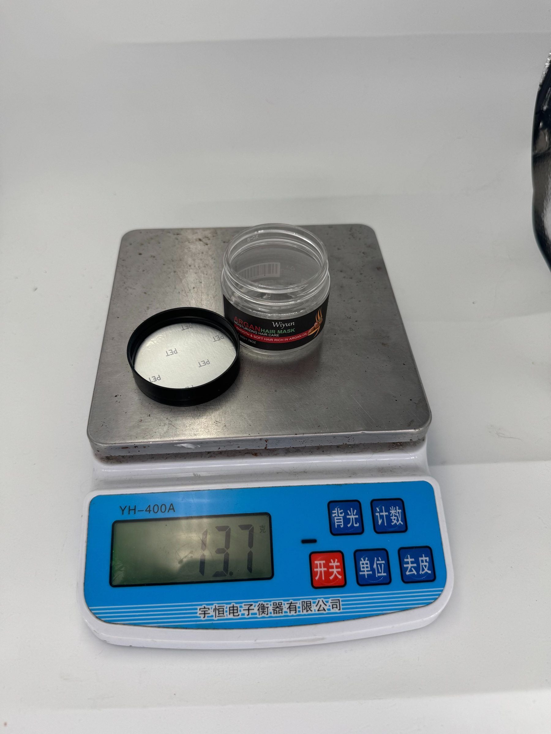A small clear plastic jar with a black lid sits on a digital scale displaying 13.7 grams.