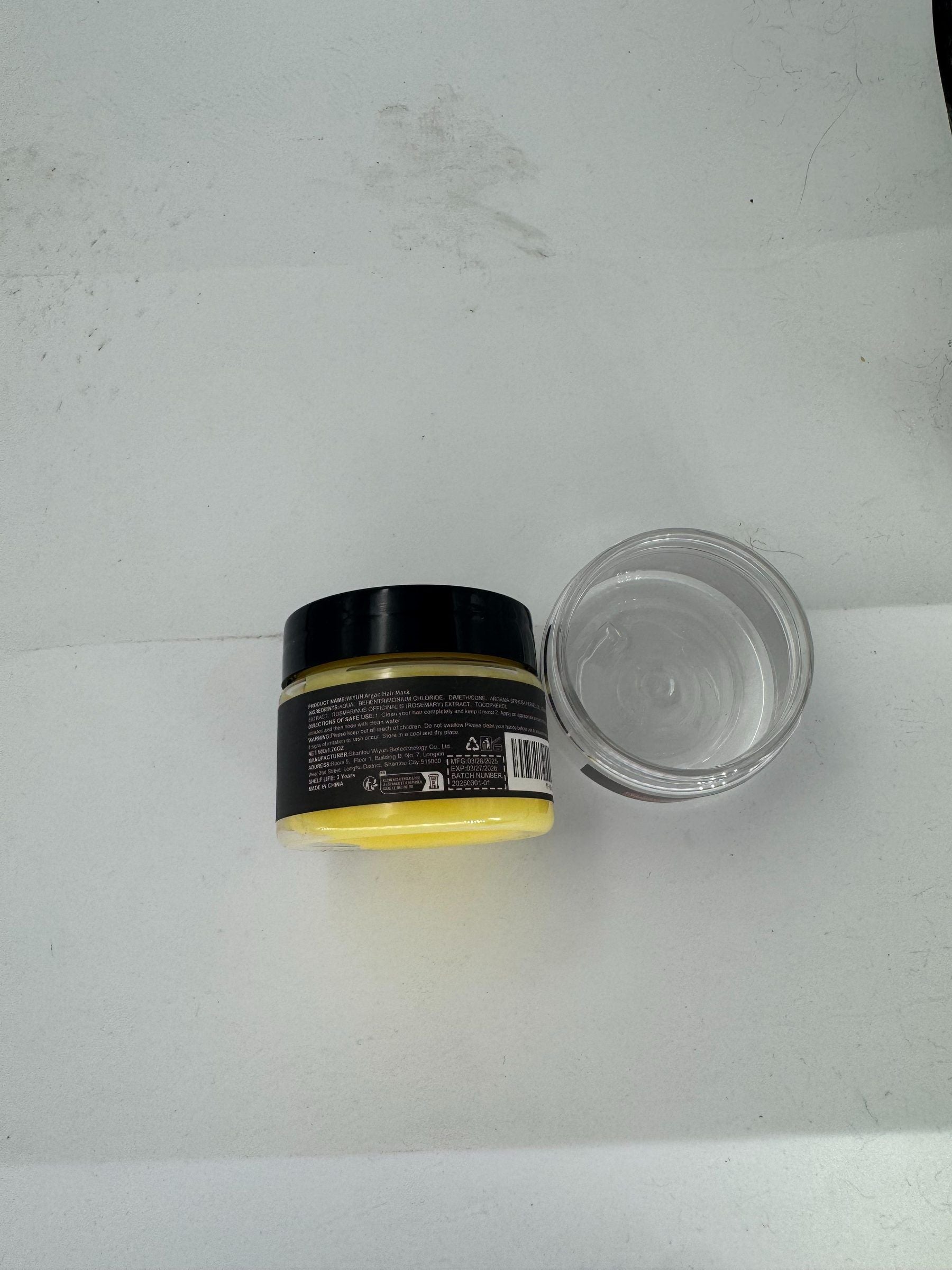 A small cosmetic jar with a black lid and yellow contents, shown next to its clear plastic lid on a white surface.