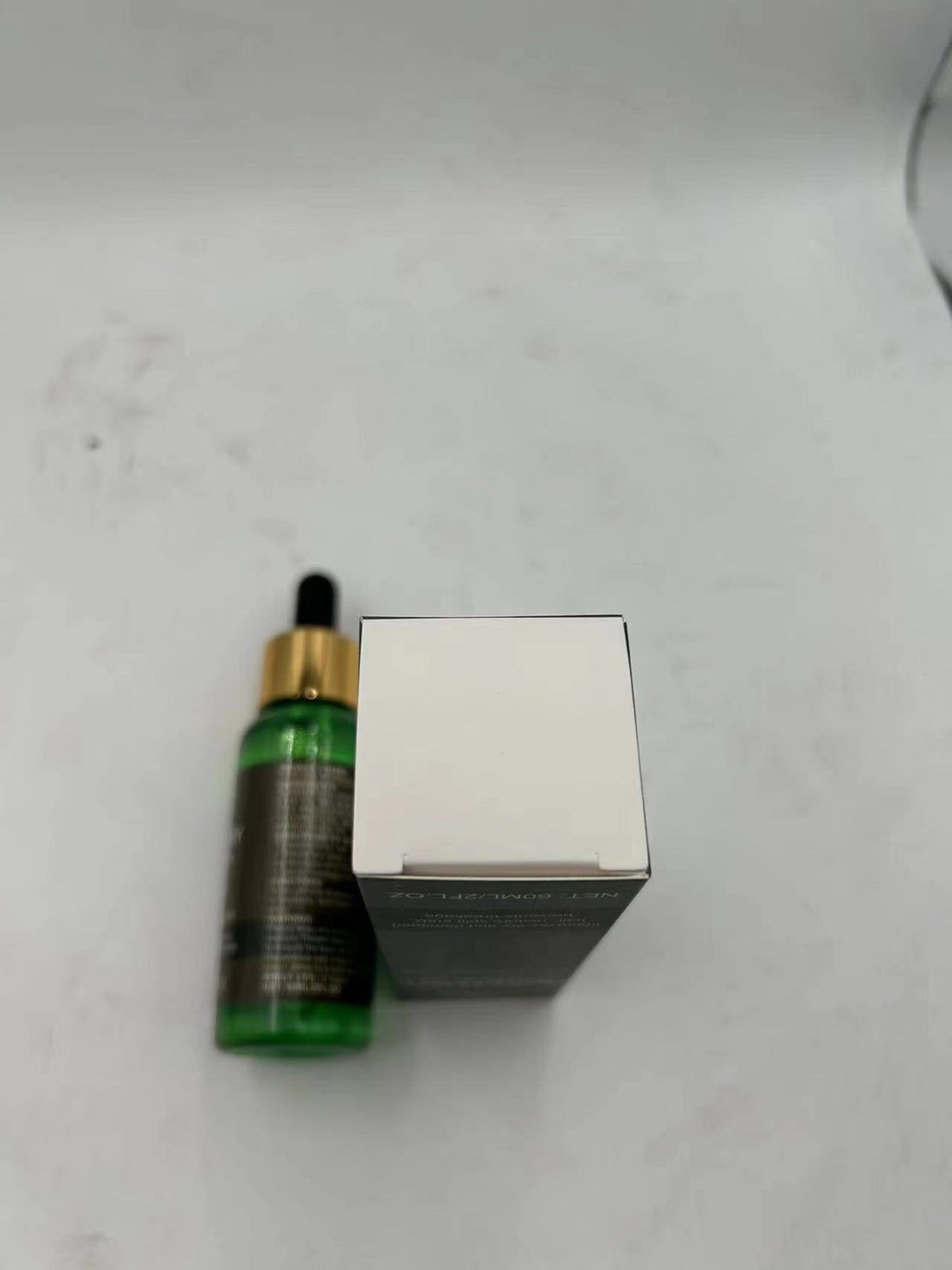 A small green glass dropper bottle with a gold collar and black cap next to its plain white box on a light gray surface.