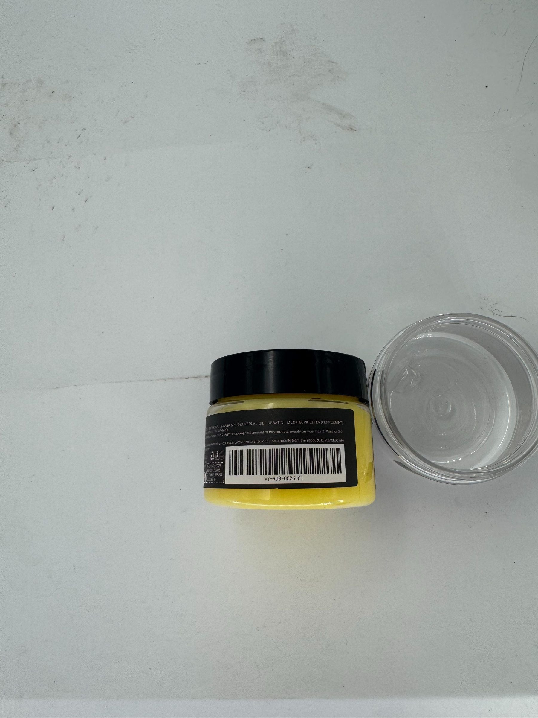 A small yellow cosmetic jar with a black lid and a black label featuring a barcode, shown next to a clear plastic lid on a light gray surface.