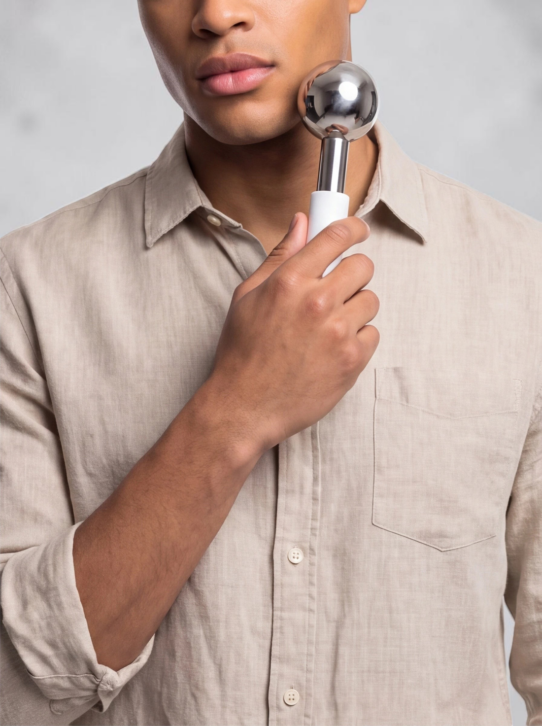 TheGromify stainless steel facial ice globe with a white handle being used to massage a man's cheek for de-puffing.
