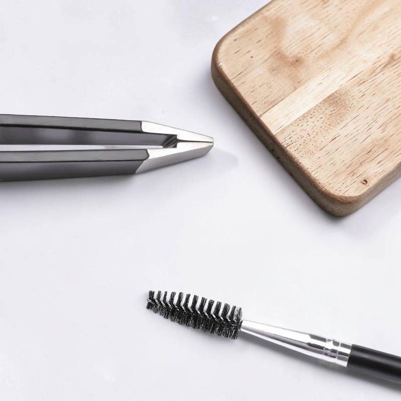 TheGromify stainless steel eyebrow tweezers and spoolie brush for precise grooming on a clean white surface