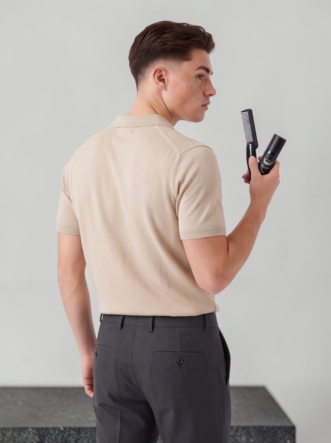 A man holding a TheGromify beard growth roller and comb, dressed in a beige polo shirt and grey trousers.